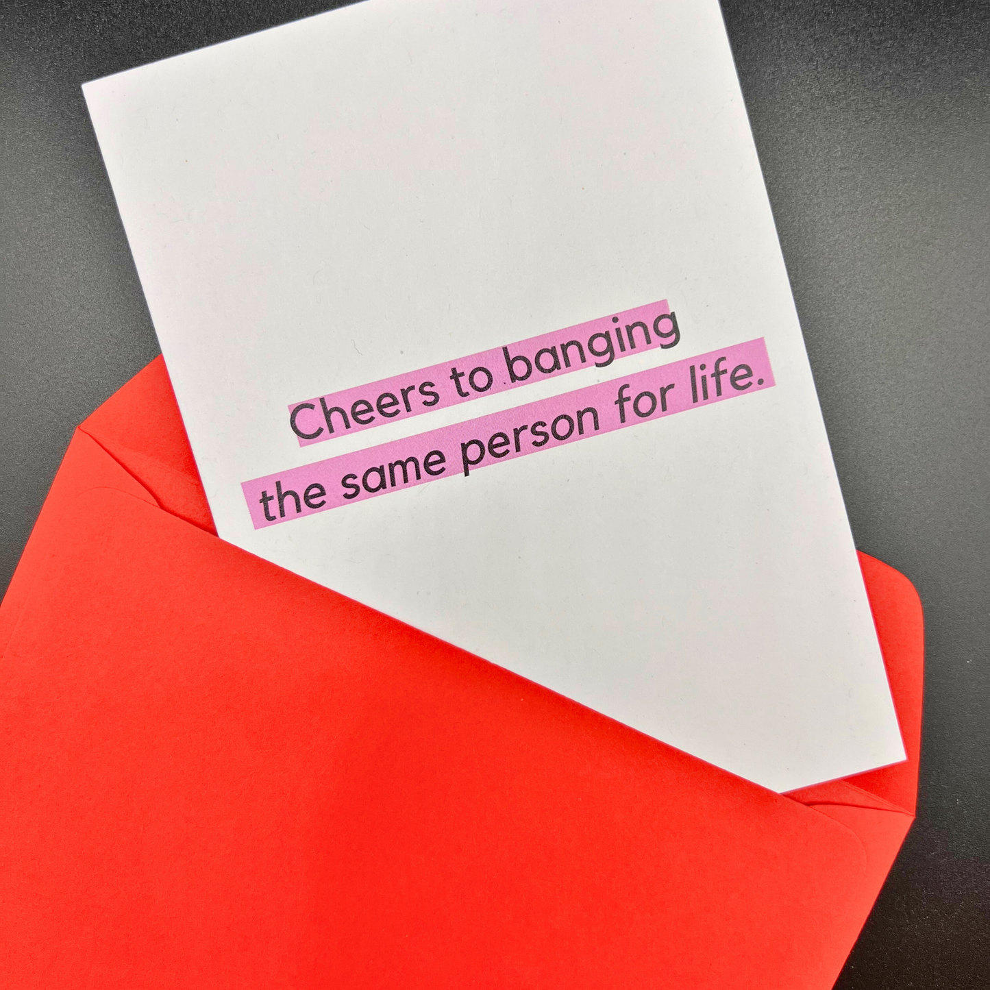 A white wedding card by The Mail Minx that says cheers to banging the same person for life. The card is slipped into a red envelope.