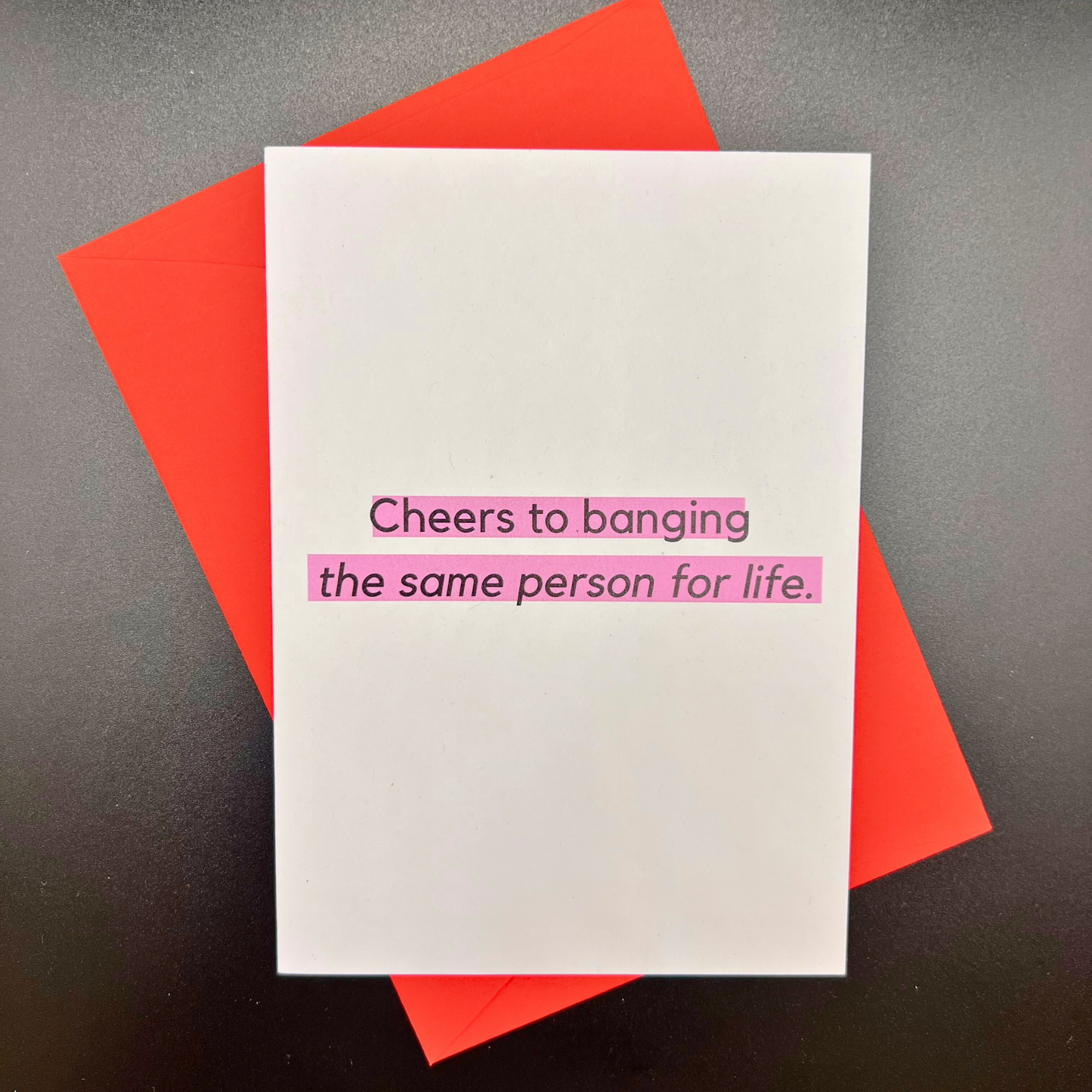 A wedding greeting card by The Mail Minx that says cheers to banging the same person for life in black text, highlighted in purple. The card has a red envelope.
