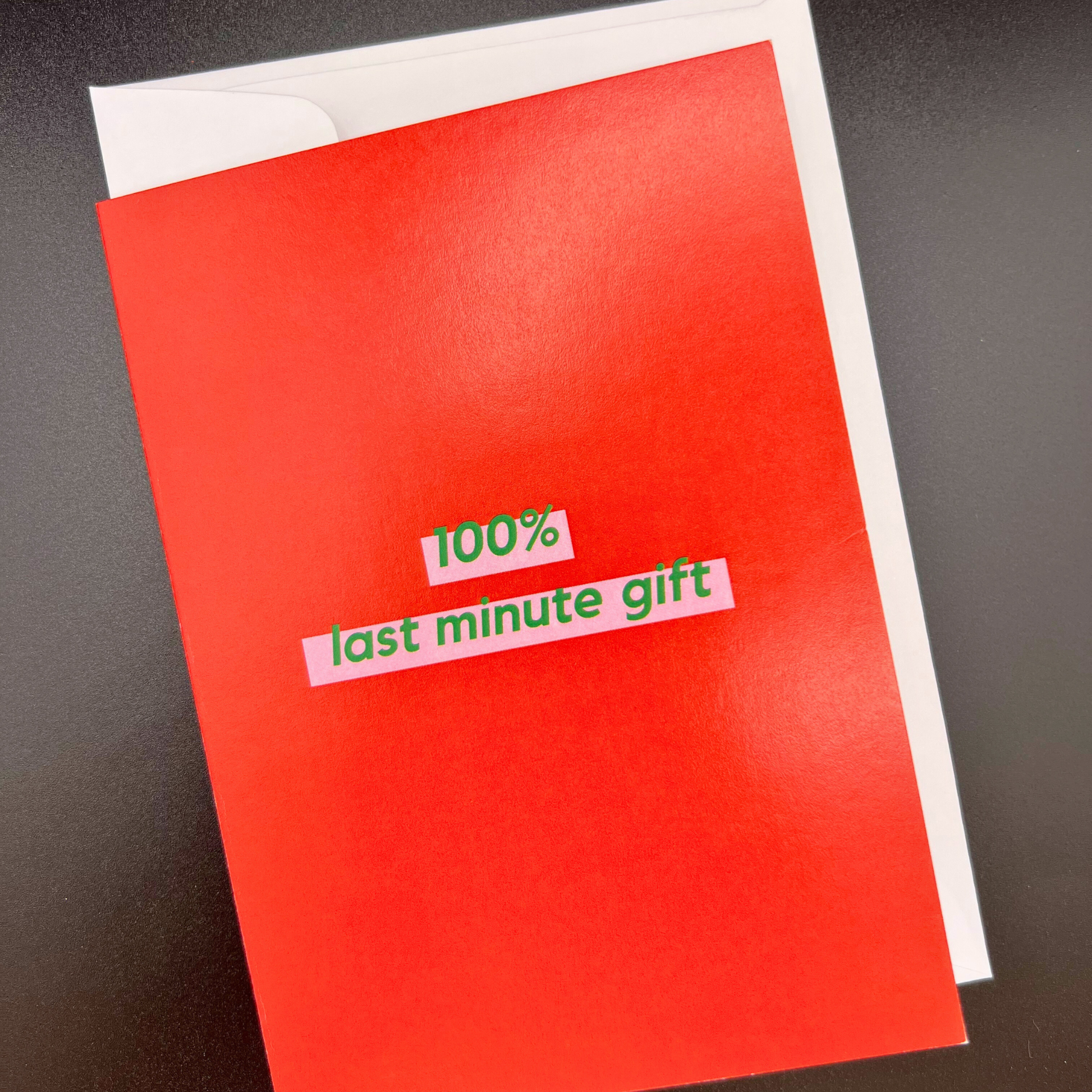 A red greeting card by The Mail Minx saying "100% last minute gift" in green highlighted in pink. The card is resting on a white envelope.