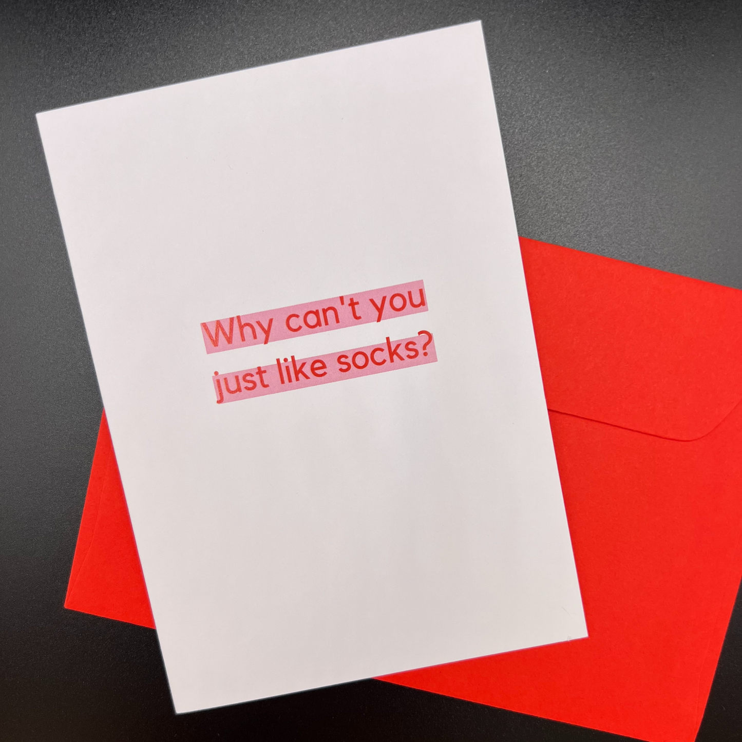 A greeting card by The Mail Minx that is white, with a message "Why can't you like socks?" in red highlighted in pink; and a red envelope.